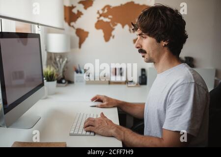 Side view of focused male freelancer sitting at table in creative workspace and typing on keyboard while working on online project Stock Photo