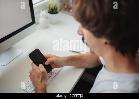From above focused male freelancer sitting at table in creative workspace using mobile phone while working on online project Stock Photo