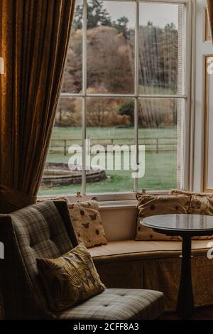 Table located near soft chair and couch with pillows against window with view of garden on autumn day Stock Photo