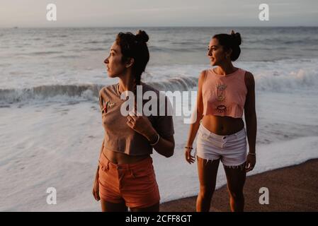 Two slim young females in shorts and bras looking away while standing on sandy shore against cloudy gray sky at sunset Stock Photo