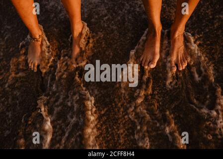 Barefoot legs of unrecognizable women standing on wet shore near splashing sea in evening Stock Photo