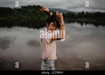 Side view of excited young female smiling and dancing with closed eyes and raised hands near pond with calm water in countryside Stock Photo