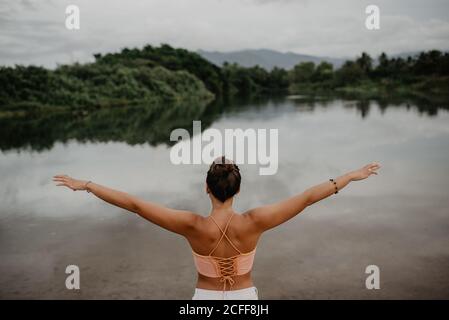 Back view of young female with outstretched arms standing near calm pond and enjoying freedom in countryside Stock Photo