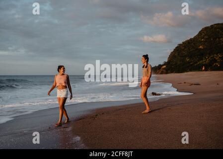 Two slim young females in shorts and bras smiling and looking at each other while standing on sandy shore against cloudy gray sky Stock Photo