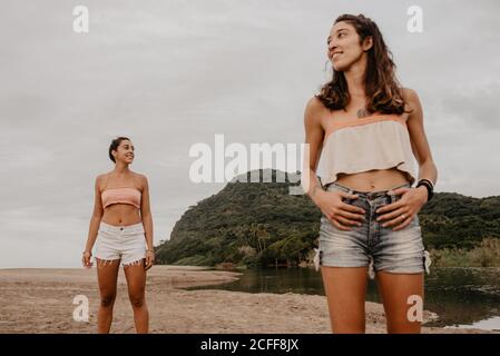 Two slim young females in shorts and bras smiling and looking at different sides while standing on sandy shore against cloudy gray sky Stock Photo