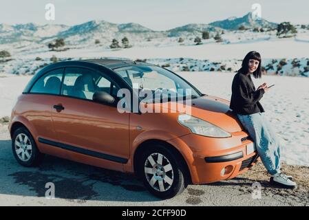Side view of young female in stylish outfit smiling and looking at camera while standing with smartphone in hands next to bright orange car on country road with snowy field and hills in background Stock Photo