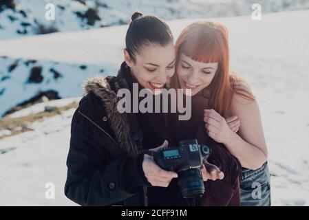 Happy female photographer in warm jacket and smiling female model looking at pictures on photo camera  during session in snowy field in sunny winter day Stock Photo
