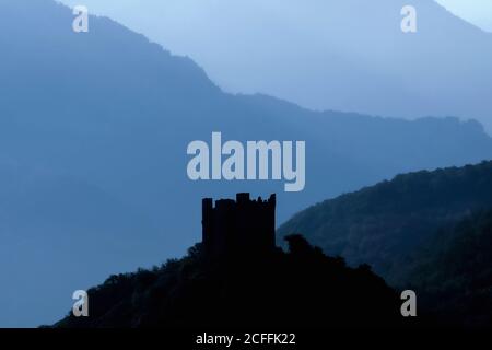 Medieval castle in the foothills of the Italian Alps.  The ruined 1300s Castello di Ussel in silhouette on a rocky hilltop at Châtillon, Valle d’Aosta, Italy.  The castle was built by the powerful Challant family and is little changed, despite being abandoned in the early 1800s and also serving time as a prison.  It was later owned by Marcel Bich (1914 - 1994), co-founder of Bic, leading ballpoint pen manufacturer.  In 1983, Baron Bich donated Ussel to the regional authority, which restored it and opened it to visitors. Stock Photo