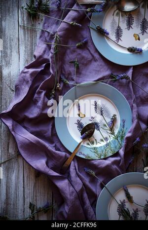 From above fresh lavenders placed around empty plates over violet fabric on lumber tabletop Stock Photo