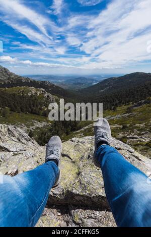 Legs of crop man sitting on stone rock above blue cloudy sky forests and mountains Stock Photo