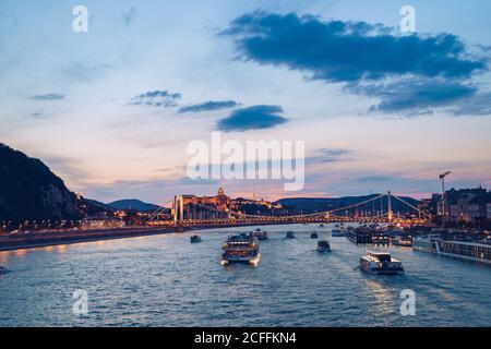 Boats on calm river reflecting colorful sunset sky and flowing under illuminated bridge in Budapest Stock Photo