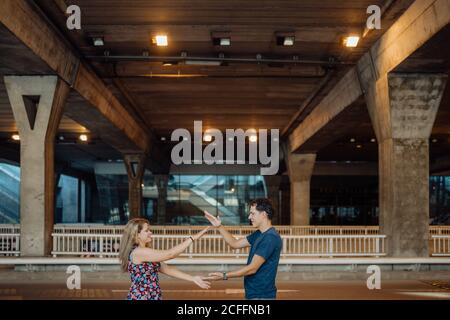 Side view of lovely couple spending time together and playing clasping hands game under large bridge in evening lights on city road Stock Photo