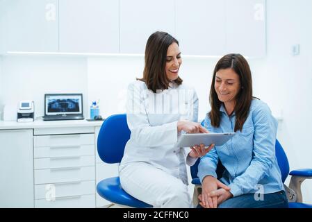 Cheerful qualified dentist and patient discussing process of treatment studying document in chair in cabinet Stock Photo