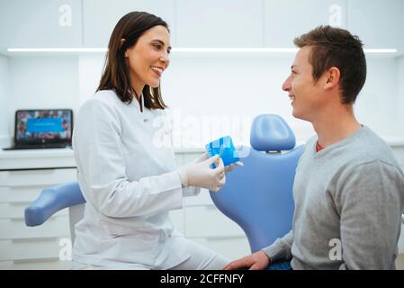 Cheerful qualified dentist and patient discussing process of treatment and doctor showing prosthesis in blue box in cabinet Stock Photo