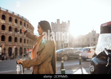 Side view of Asian female with suitcase smiling and stretching out arm while hailing cab on street of tourist city Stock Photo