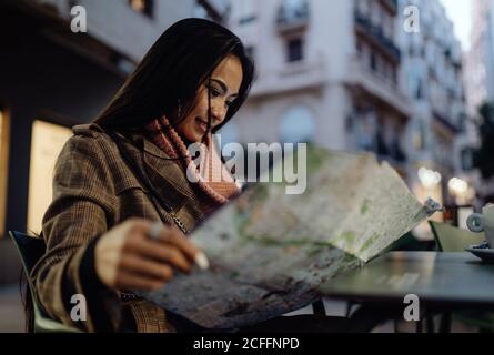Young Asian Woman smiling and reading map while sitting at table in street cafe in evening in city Stock Photo