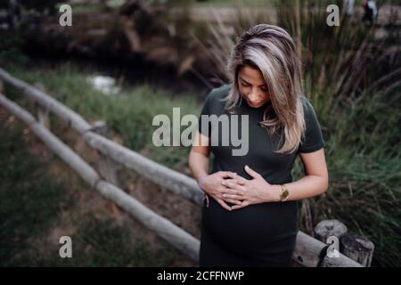 Beautiful pregnant female in casual dress smiling and holding hands on belly while standing next to wooden bridge over small river in park in summer day Stock Photo