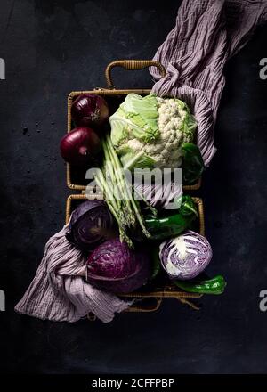 Top view of various fresh ripe vegetables with violet towel in wicker basket placed on dark surface as concept of healthy food Stock Photo