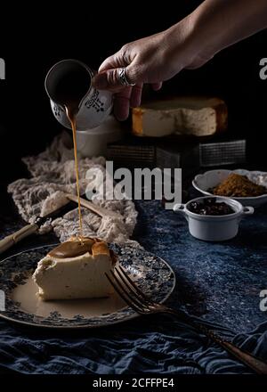 Faceless female cook pouring caramel on piece of appetizing cheese pie on ceramic plate placed on blue table against black background after cooking dessert at home Stock Photo