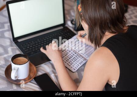 From above back view of crop female in casual outfit lying on bed with laptop and cup of coffee while writing in notepad and relaxing during weekend Stock Photo