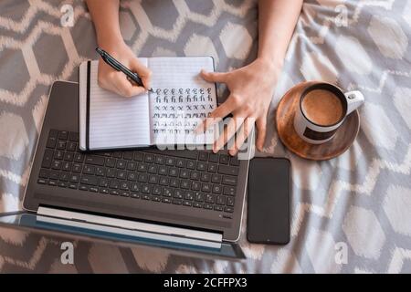 Top view of crop Woman taking notes in bed while working next to his laptop, smartphone and cup of coffee Stock Photo