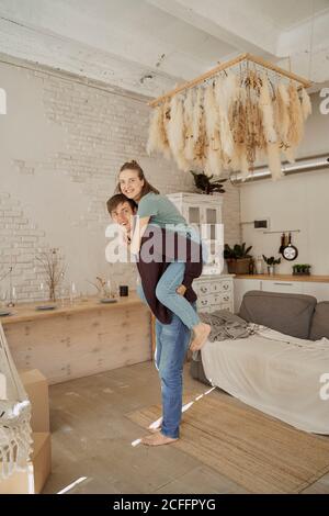 Side view of smiling handsome guy giving piggyback ride to cheerful female in light cozy living room Stock Photo