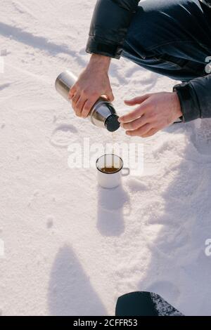 Crop male traveler pouring hot tea from thermos bottle to enamel mug placed on snow during winter trip in Siberia Russia Stock Photo