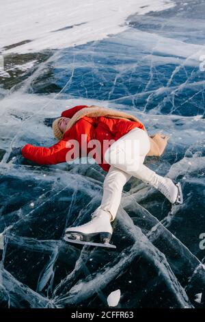 From above of happy young active female in warm active wear and skates lying on ice on frozen river and enjoying nice winter day in Siberia Stock Photo