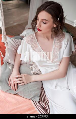 Gorgeous Woman with red lips in white dress looking away while sitting on floor beside sofa Stock Photo