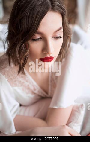 Gorgeous Woman with red lips in white dress looking away while sitting on floor beside sofa Stock Photo