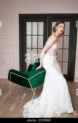 Barefooted slender bride in elegant white dress looking at camera while sitting on backrest of green armchair against black doors in spacious apartment Stock Photo