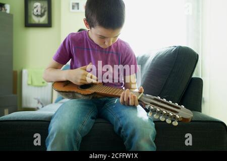 Focused boy in casual jeans and shirt leaning to play ukulele sitting on comfortable sofa in light living room at home Stock Photo