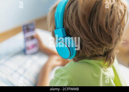 Back view boy in casual wear with headphones lying on bed and watching movie on tablet while resting at home Stock Photo