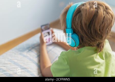Back view boy in casual wear with headphones lying on bed and watching movie on tablet while resting at home Stock Photo