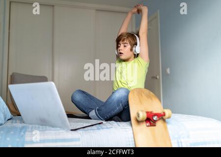 Depressed little boy with headphones on neck sitting on bed and using laptop while spending time during self isolation because of coronavirus at home Stock Photo