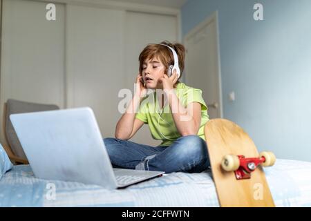 Depressed little boy with headphones on neck sitting on bed and using laptop while spending time during self isolation because of coronavirus at home Stock Photo