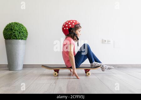 Little kid in red spotted helmet and casual clothes and dreaming about outdoors amusement while sitting alone on longboard in corridor of light modern apartment staying at home Stock Photo
