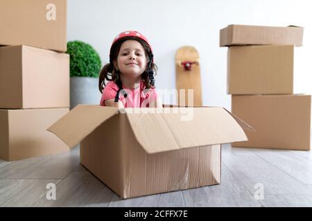 Happy little girl smiling and fastening helmet while sitting in cardboard box and playing during relocation Stock Photo
