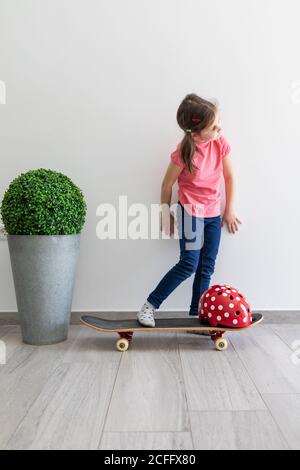 Kid in casual clothes with longboard and vivid red protective helmet in light contemporary flat dreaming about street staying at home Stock Photo
