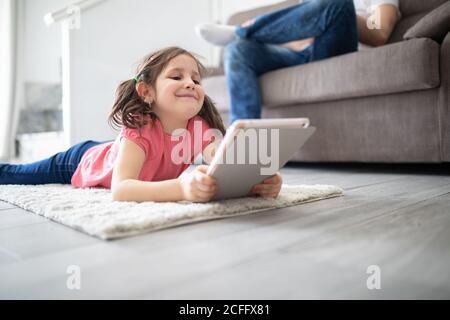Joyful small kid focusing on screen with interest while lying on carpet on floor and watching cartoon using digital tablet in light living room of modern apartment Stock Photo