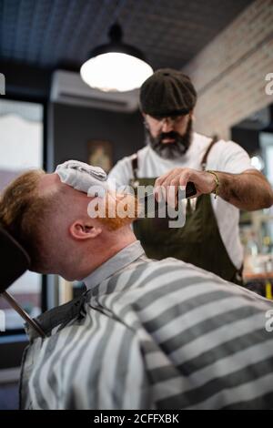 Blurred unrecognizable barber with comb fixing redhead man beard with towel covering eyes sitting in barbershop Stock Photo