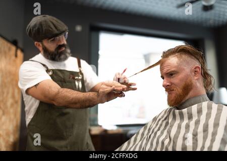 Modern hairdresser barber cutting a adult man redhead's hair in barber chair