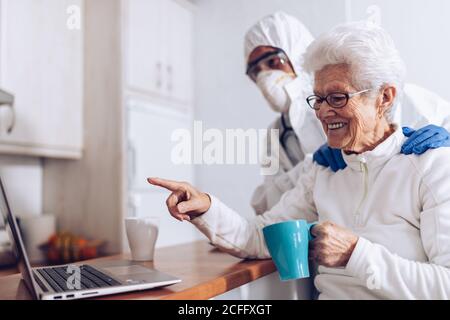 Cheerful old female drinking tea and chatting with friend during video call via laptop while home care specialist in protective costume and mask standing nearby during coronavirus lockdown Stock Photo