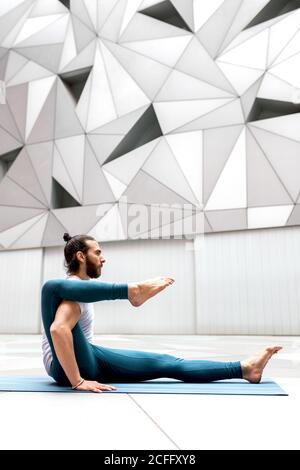 Side view of adult bearded man keeping leg on shoulder while doing stretching yoga exercise against wall with geometric ornament Stock Photo