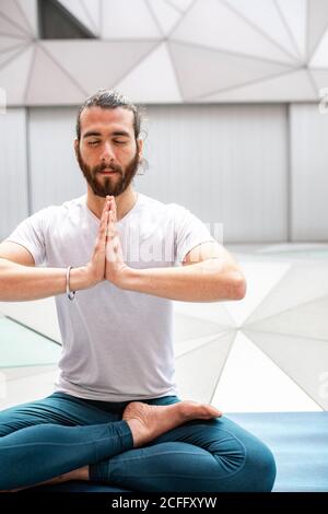 Adult bearded man in sportswear sitting crossed legged and meditating with closed eyes and clasped hands during yoga training in geometric room Stock Photo