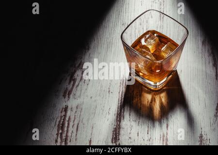 From above transparent square shaped glass of whiskey drink with ice cubes on shabby wooden table in rays in dark room Stock Photo