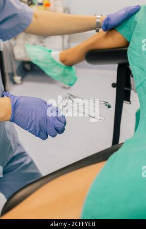 Faceless female doctor in blue gloves and uniform with plastic gynecological mirror examining anonymous patient in shoe covers lying on chair in fertility clinic Stock Photo