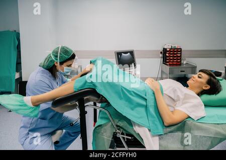 Side view of female doctor in blue uniform and sterile mask examining smiling patient on gynecological chair in fertility clinic Stock Photo