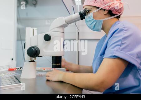 Side view of Woman in medical uniform and mask using modern microscope to examine human cells while working in lab of modern clinic Stock Photo