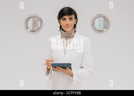 Positive medical practitioner using tablet and looking at camera while standing against white door in hallway of modern hospital Stock Photo